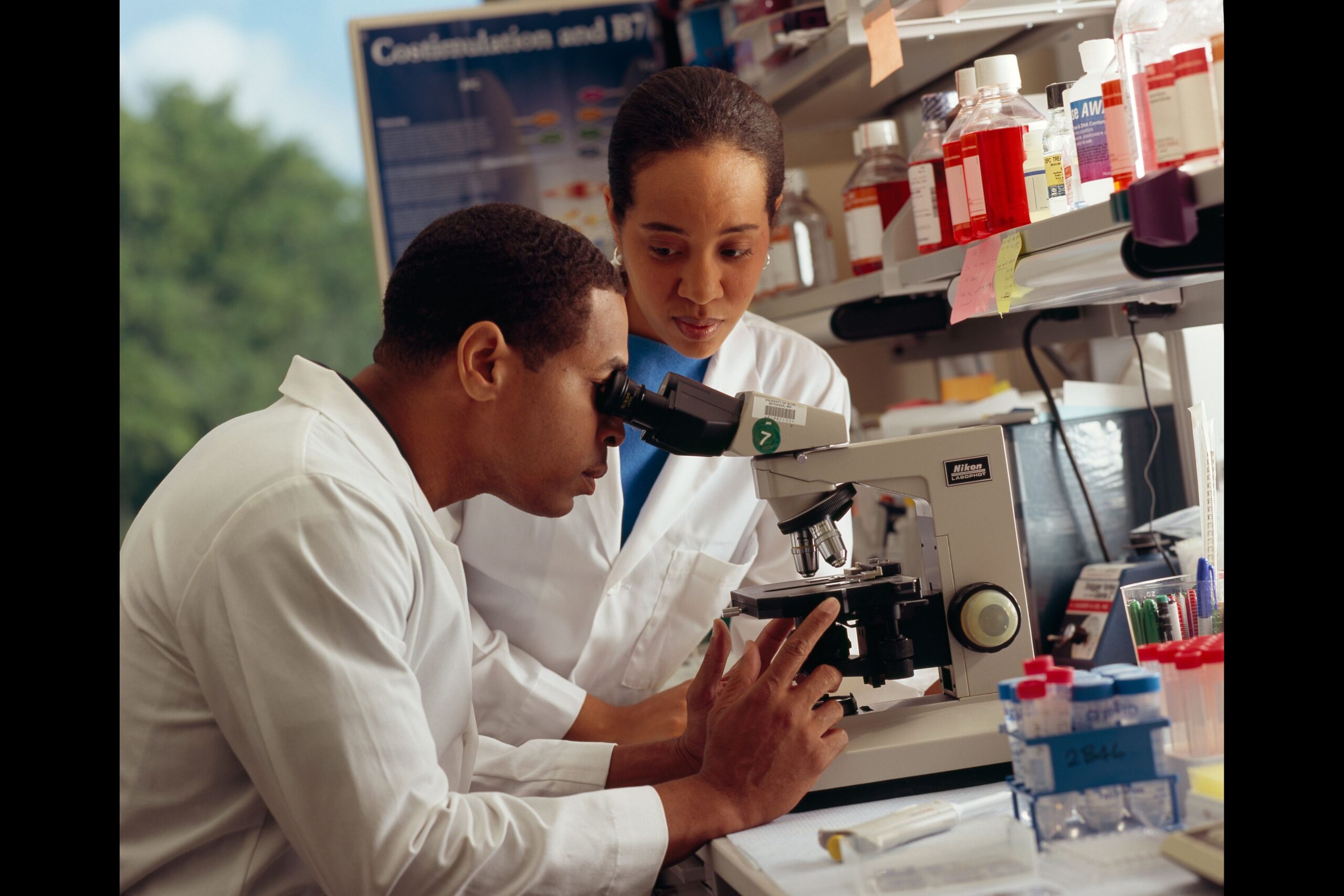 Deux chercheurs en sarrau blanc observent au microscope, dans un laboratoire — National Cancer Institute, domaine public, via Wikimedia Commons
